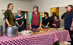 Five women and one girl stand in discussion at a cloth covered table filled with pots, vegetables and cutting boards